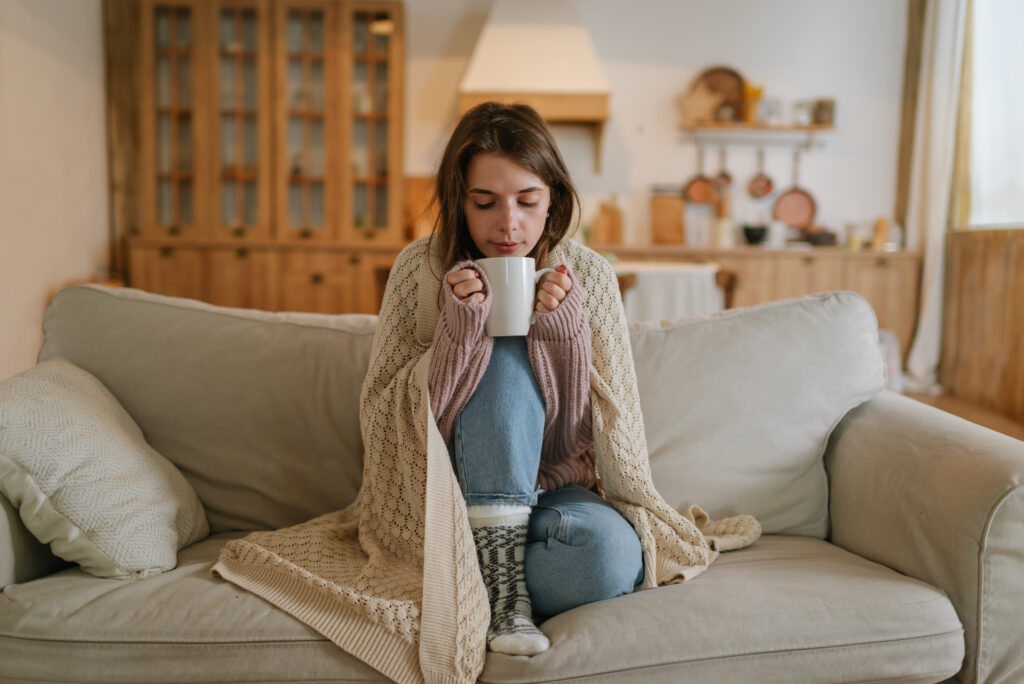 A young woman sits cross-legged on a sofa, wrapped in a cozy blanket, holding a white mug with both hands. She appears relaxed and warm in a softly lit, homey living room setting.