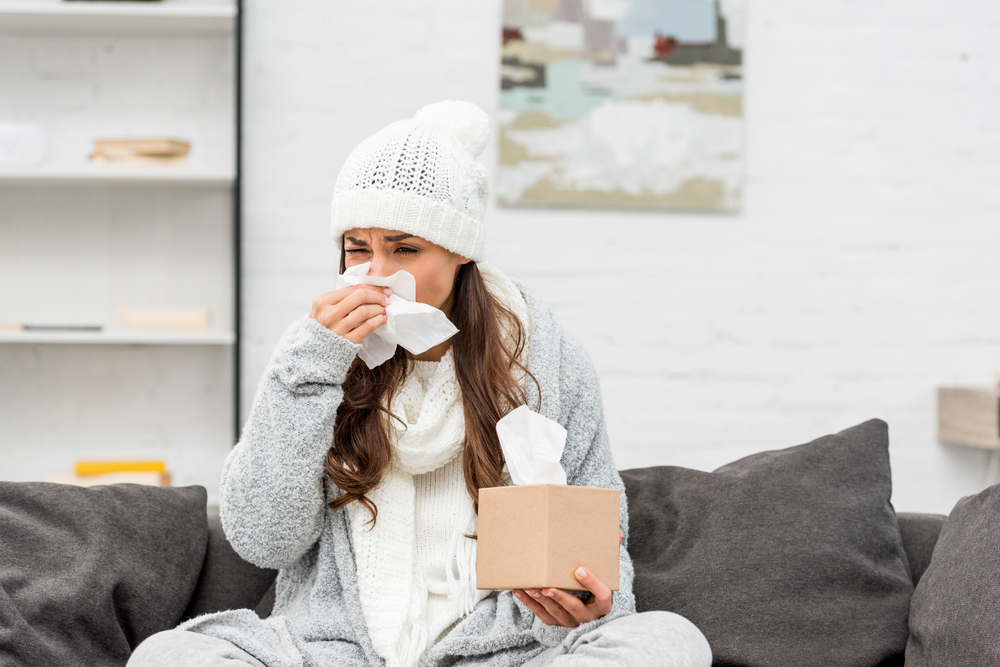 A woman wearing a white knit hat and scarf sits on a couch, holding a tissue box and using a tissue to blow her nose, appearing to have a cold. The background shows shelves and a painting.