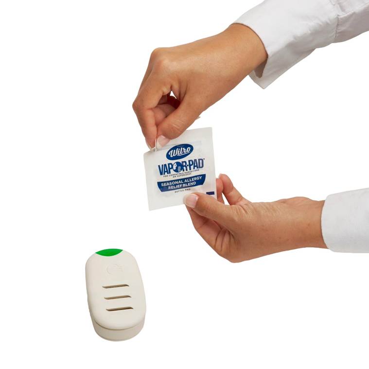 A person’s hands are opening a Vicks VapoPad packet above a white vaporizer device with a green button, set against a plain white background.